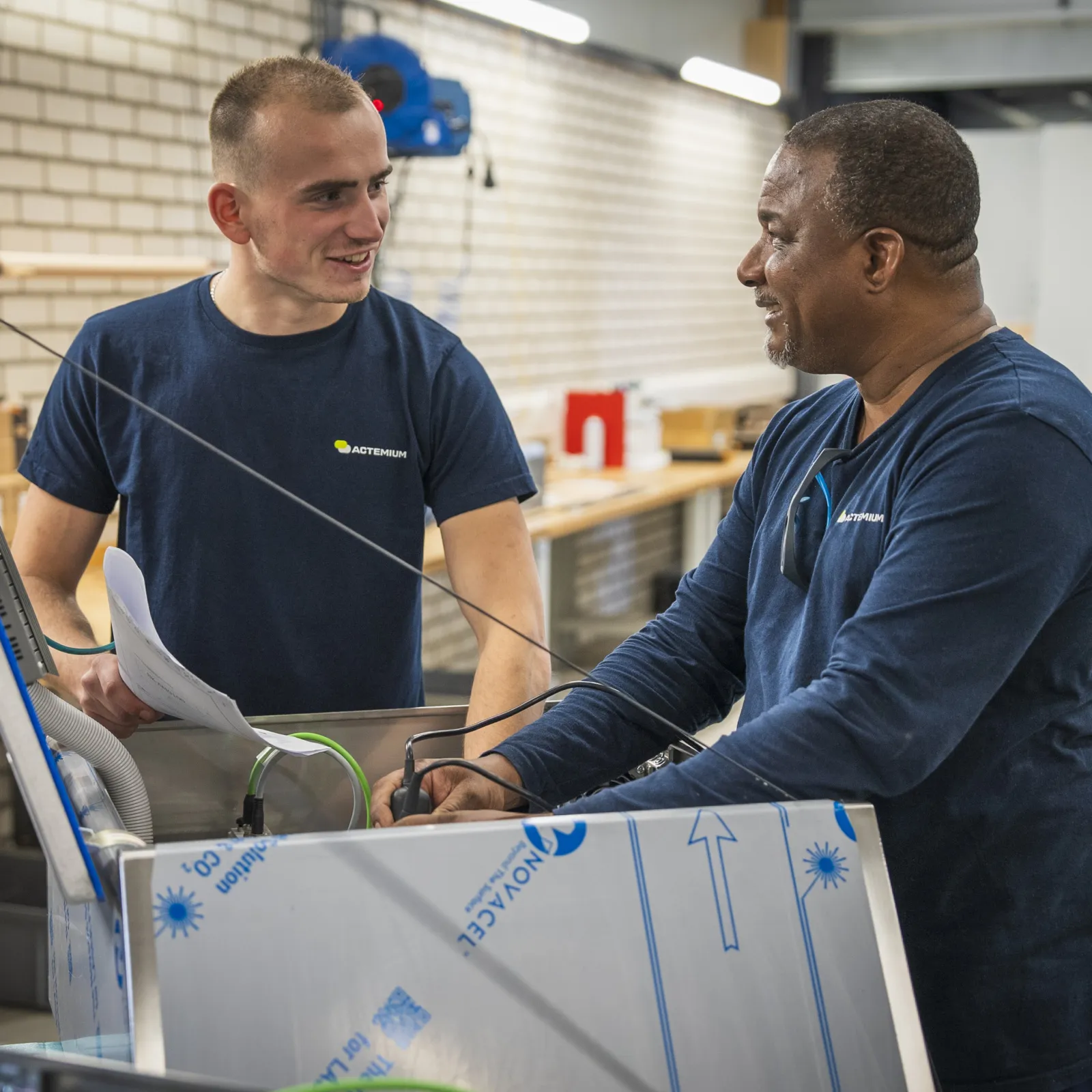 Twee collega's van Actemium werken samen aan een technische installatie in een werkplaats. Op een scherm worden instellingen en technische gegevens bekeken, terwijl één collega documentatie vasthoudt en de andere collega de installatie bedient. De foto laat de samenwerking, kennisdeling en praktijkgerichte werkomgeving binnen Actemium zien, waar collega's samen werken aan technische oplossingen en installaties.